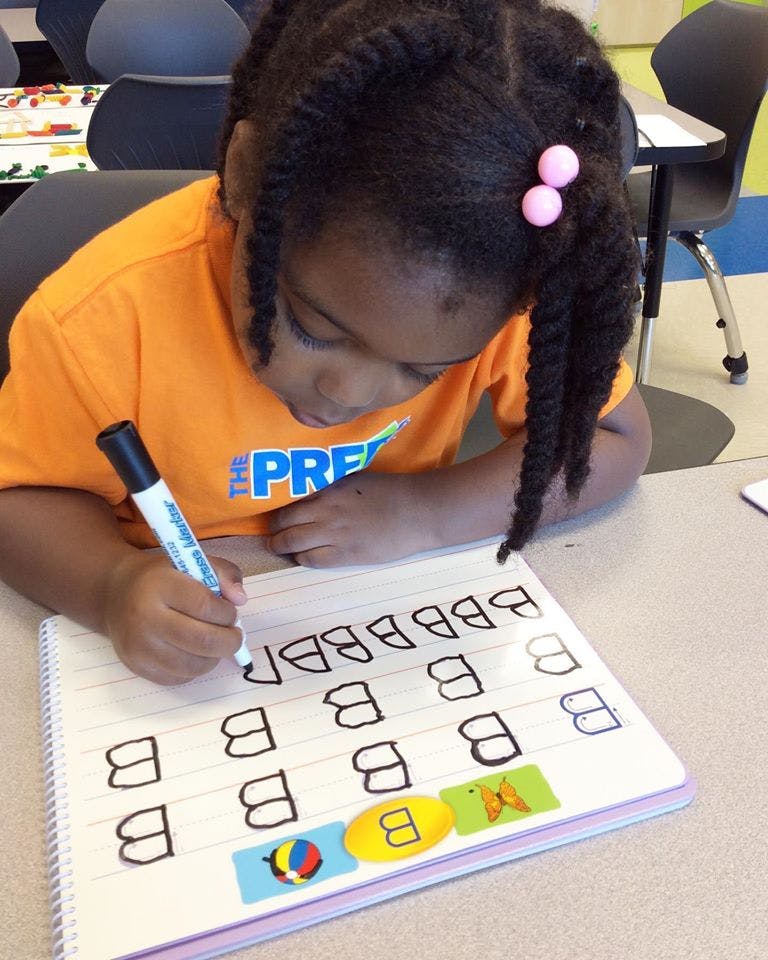 Child writing the letter B on a notebook with a marker in a classroom