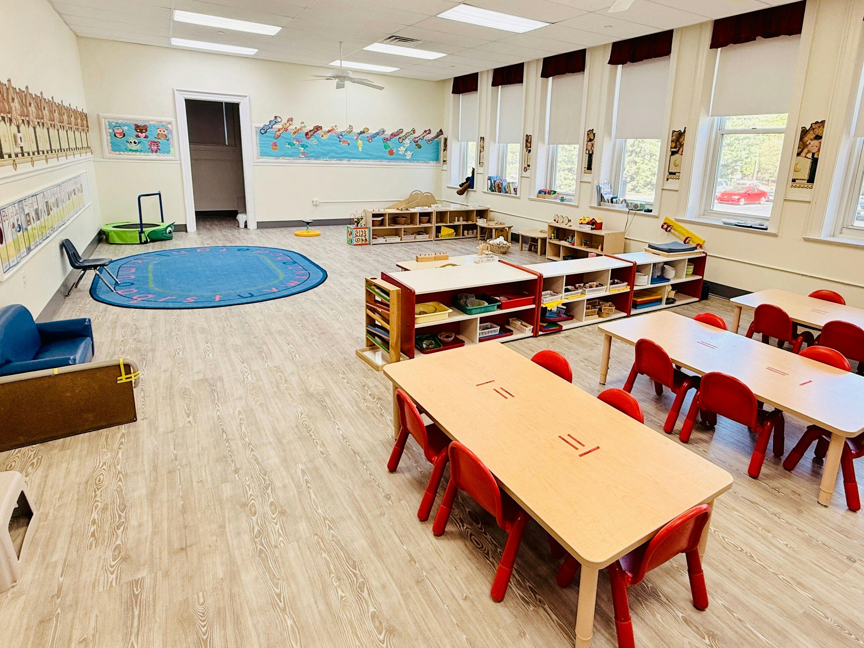 Classroom with small tables and chairs, shelves with toys, and a blue rug