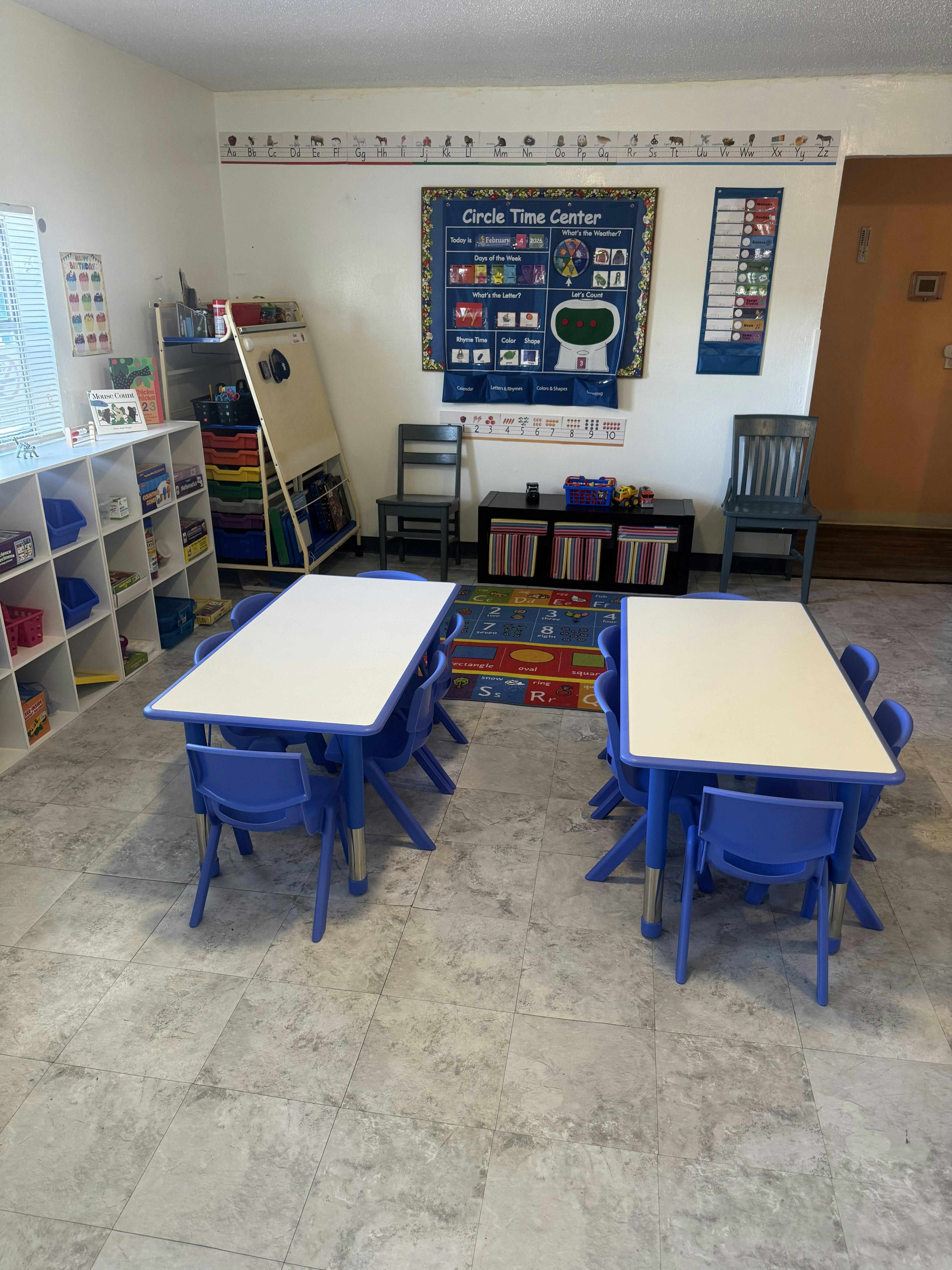 Classroom with two blue tables and chairs, white shelving unit, and learning posters