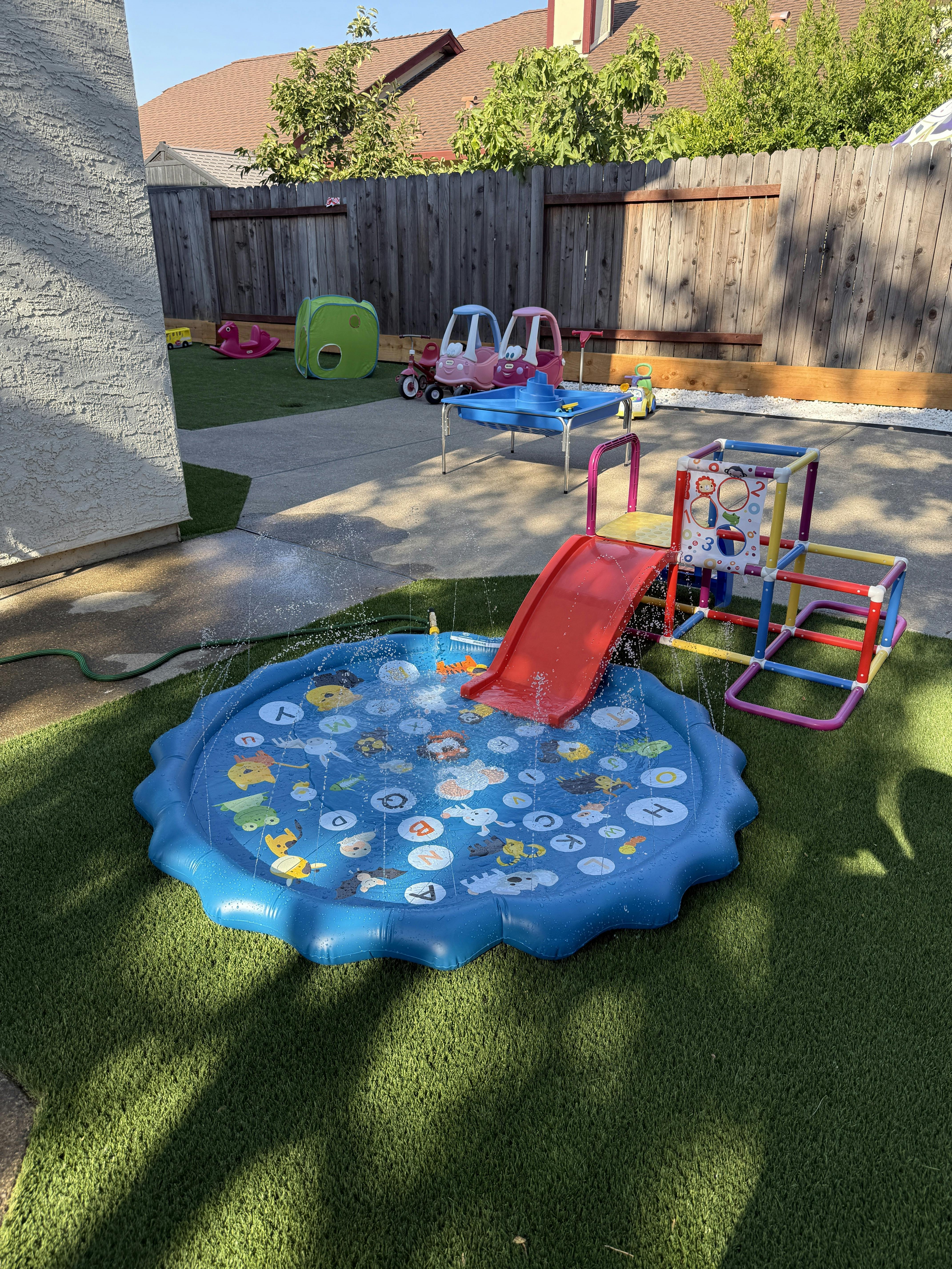 backyard play area with a blue splash pad and red slide, artificial turf
