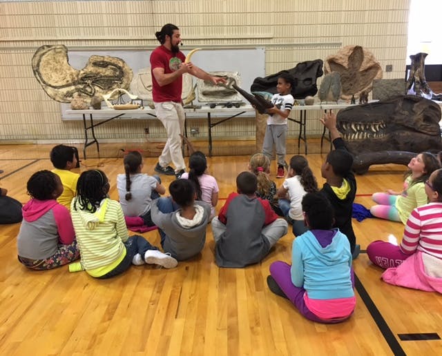 Person presenting dinosaur fossils to children sitting on a gym floor