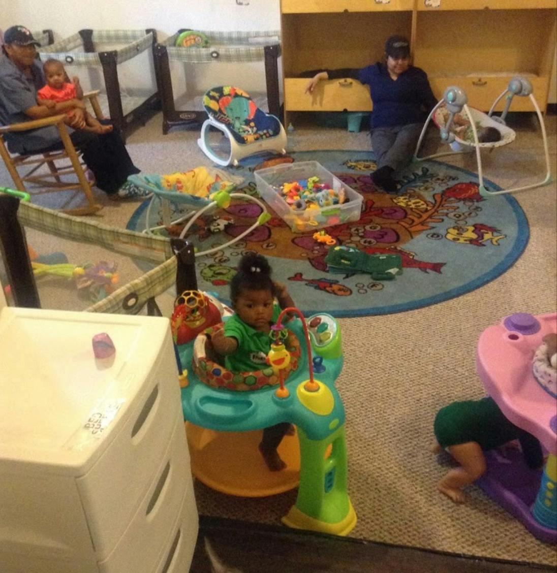People and children in a room with play equipment and cribs