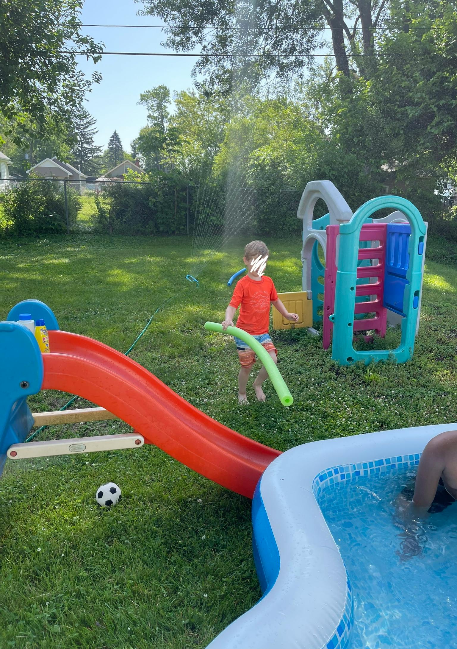 Child playing with a pool noodle in a backyard with a slide and inflatable pool