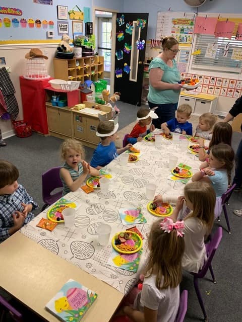 Adult serving food to children seated at a long table in a classroom