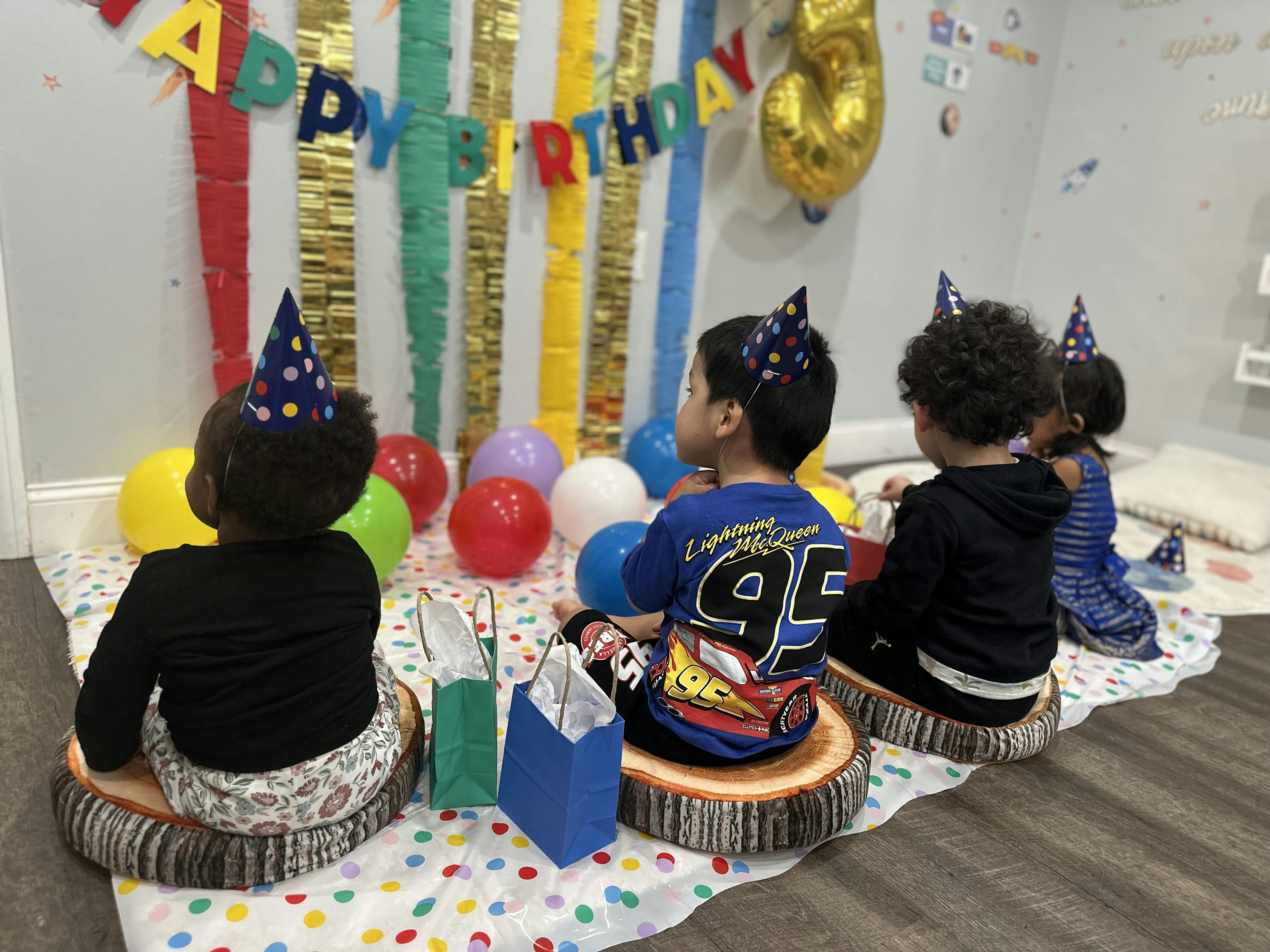 Children in party hats sitting on the floor with balloons and "HAPPY BIRTHDAY" banner