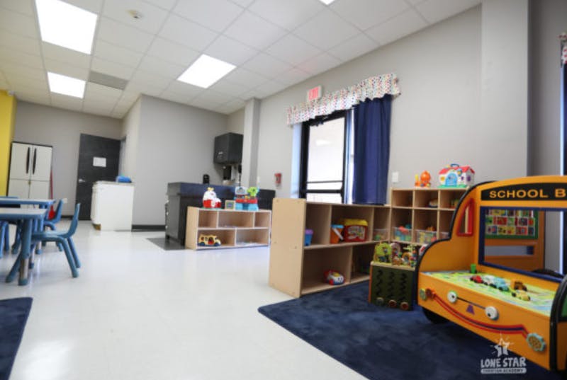 Empty classroom with small tables and chairs, shelves with toys, and a toy school bus