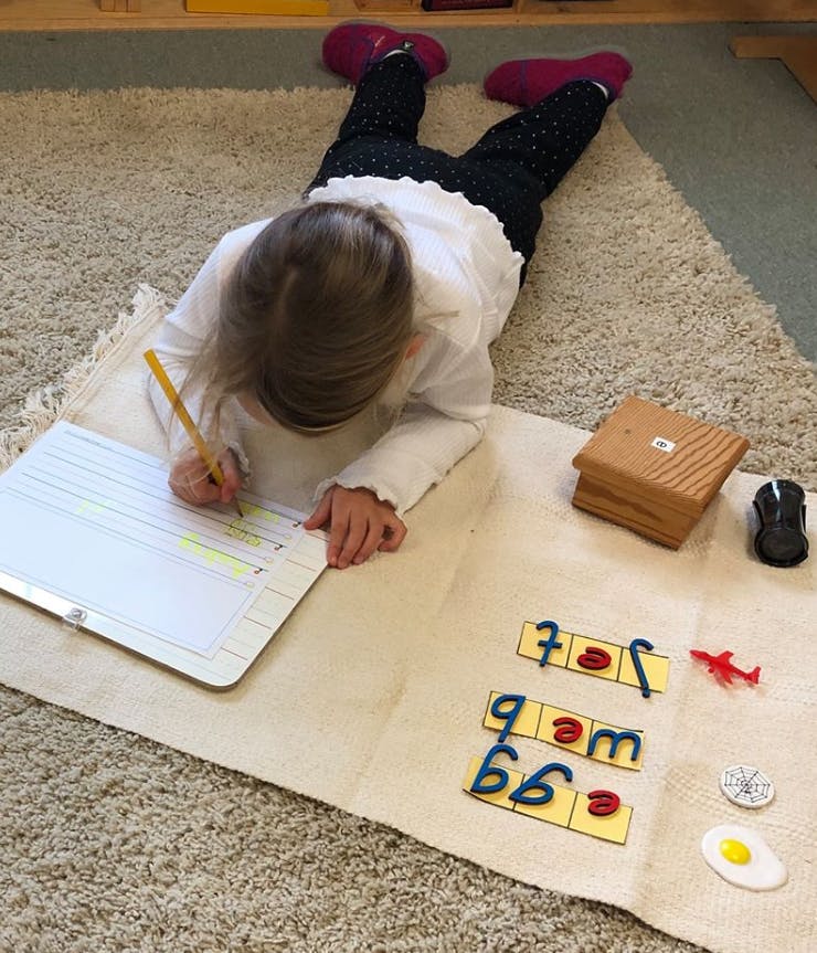 Child lying on floor writing on paper with pencil, learning materials nearby