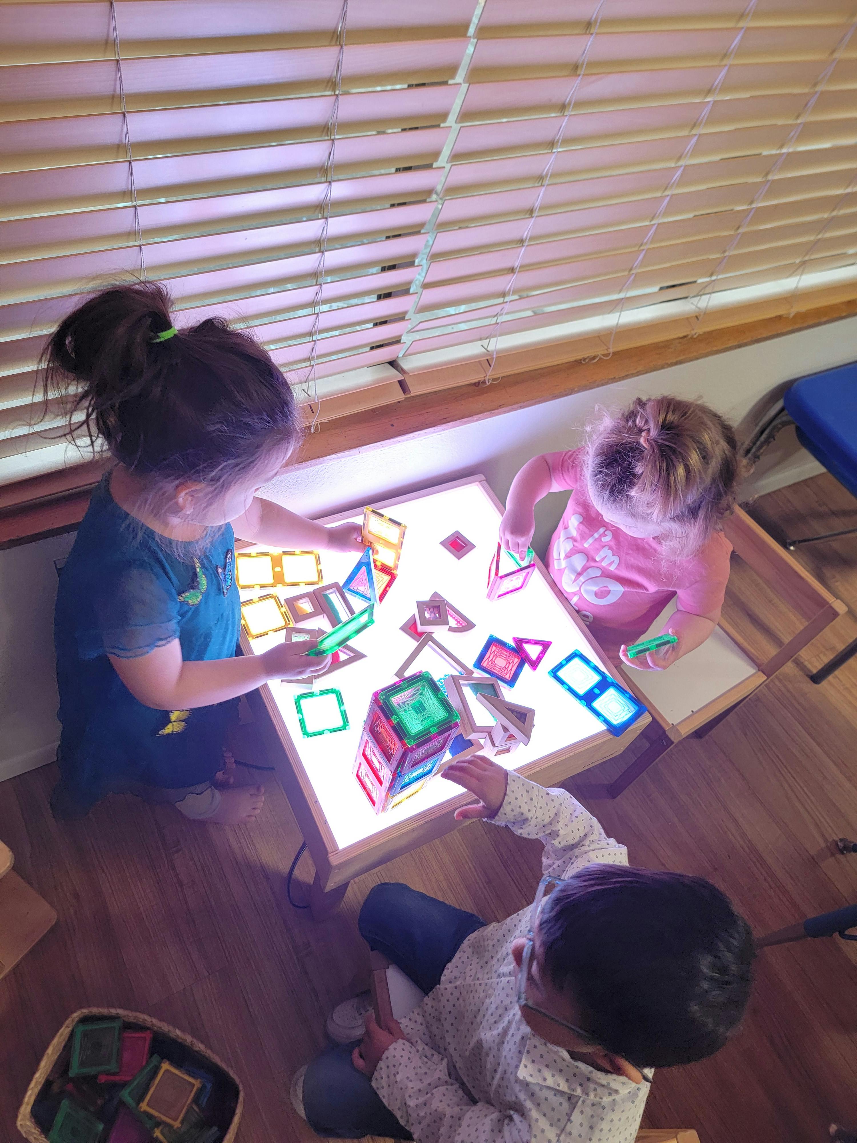 Three children building with colorful magnetic tiles on a light table