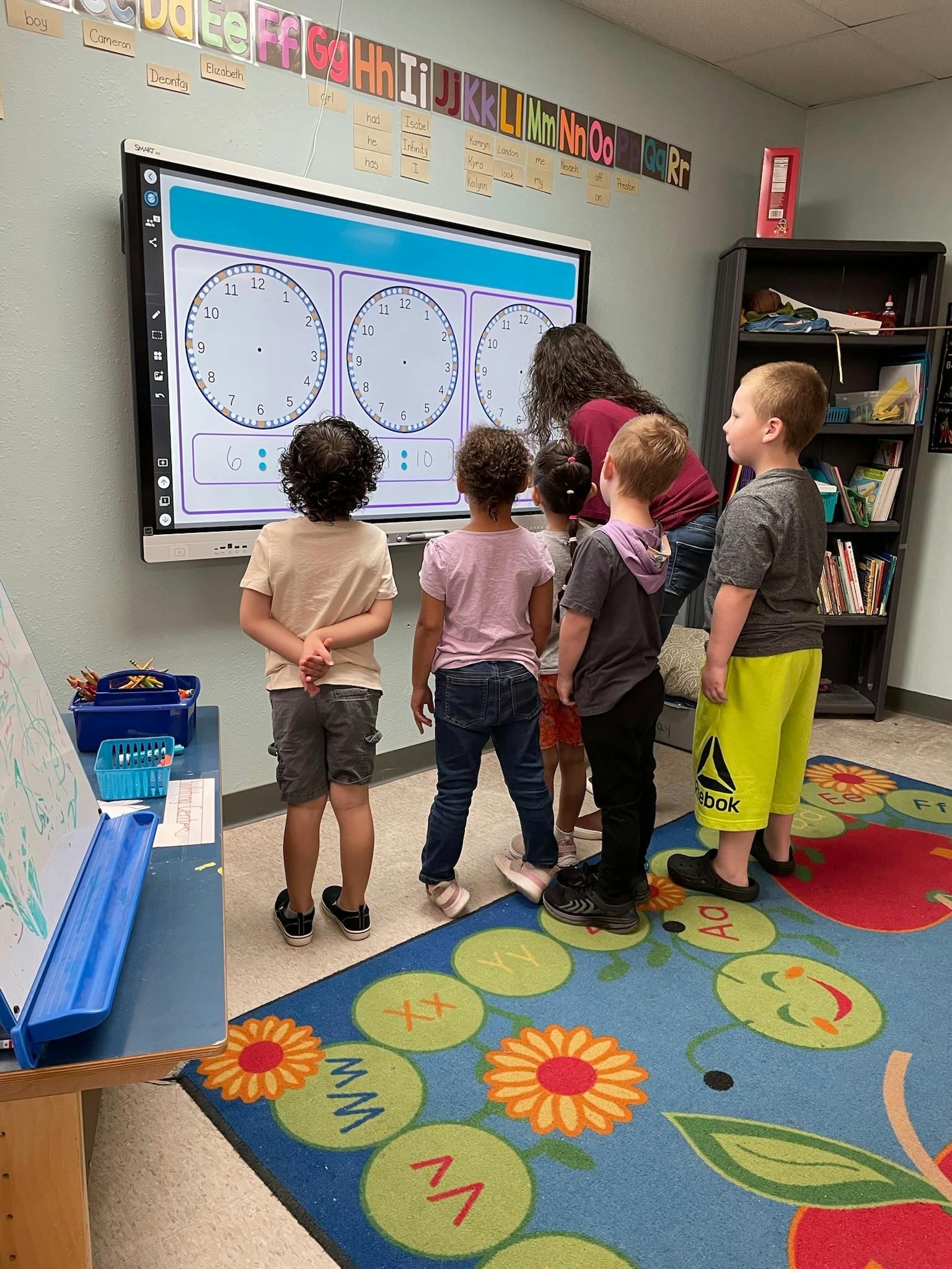 Adult and children looking at a large screen displaying clock faces in a classroom