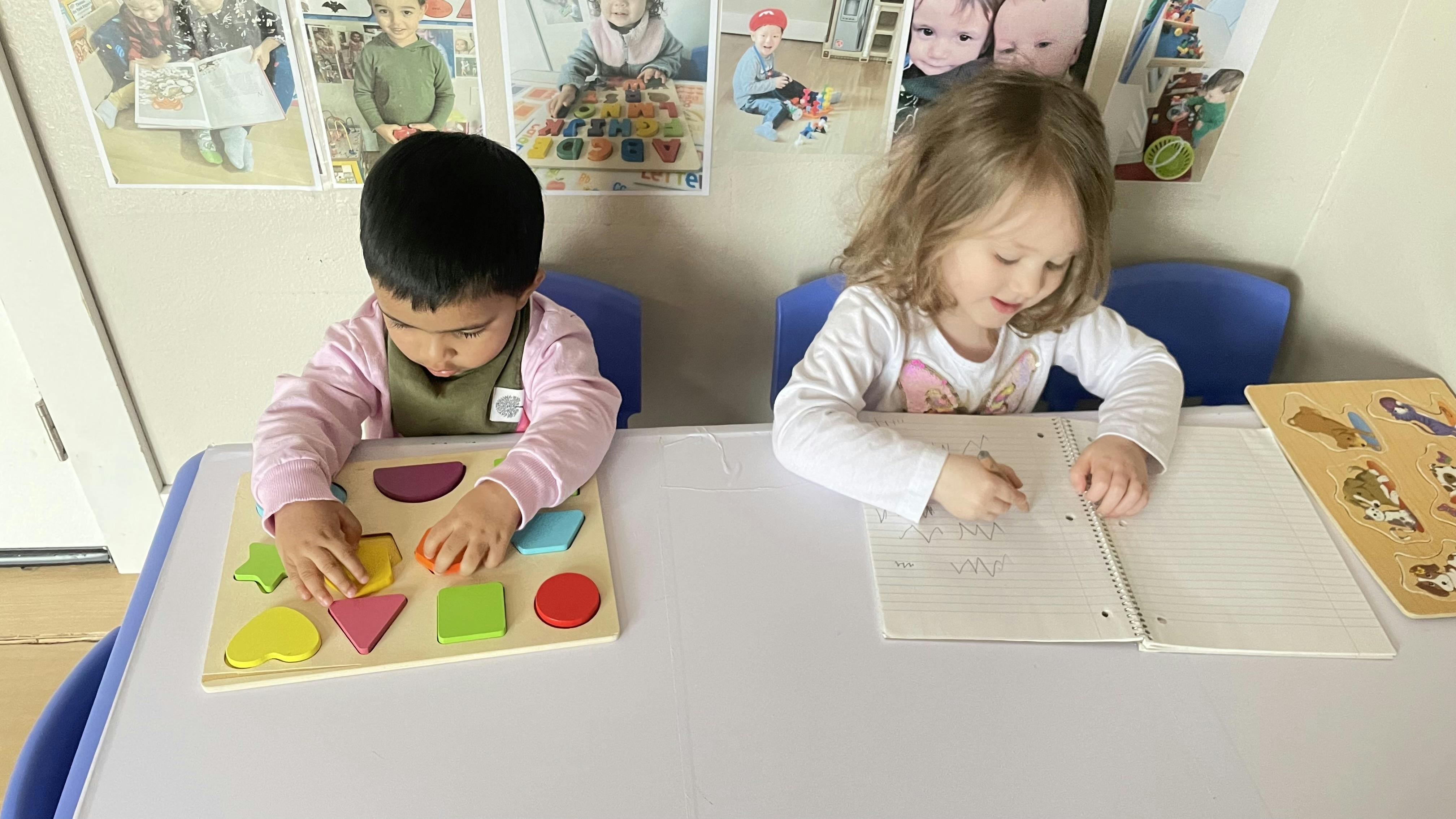 Two children at a table, one playing with a shape puzzle, one drawing