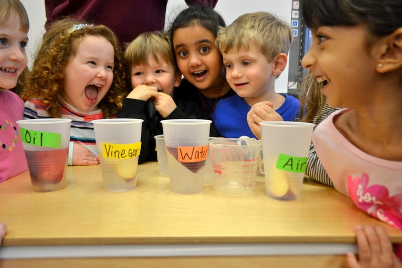 Children gathered around a table with cups labeled "Oil," "Vinegar," "Water," and "Air"