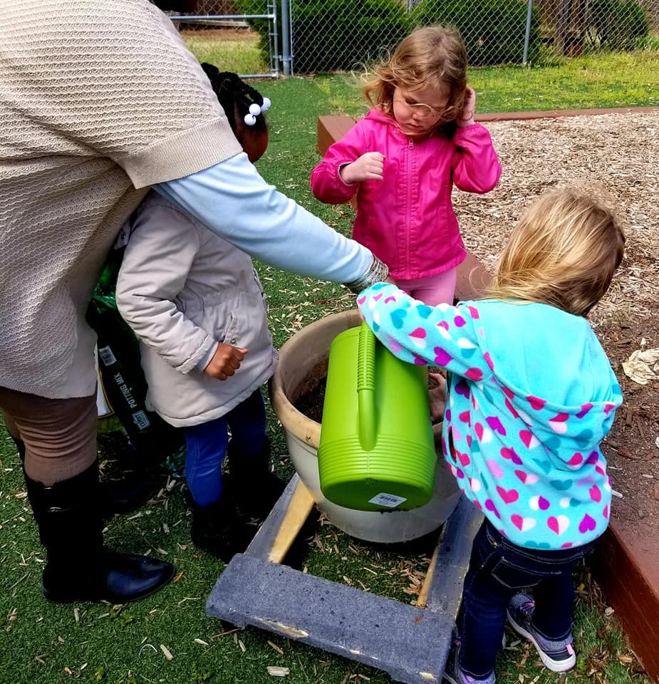 Adult and three children gathered around a large pot with a green watering can