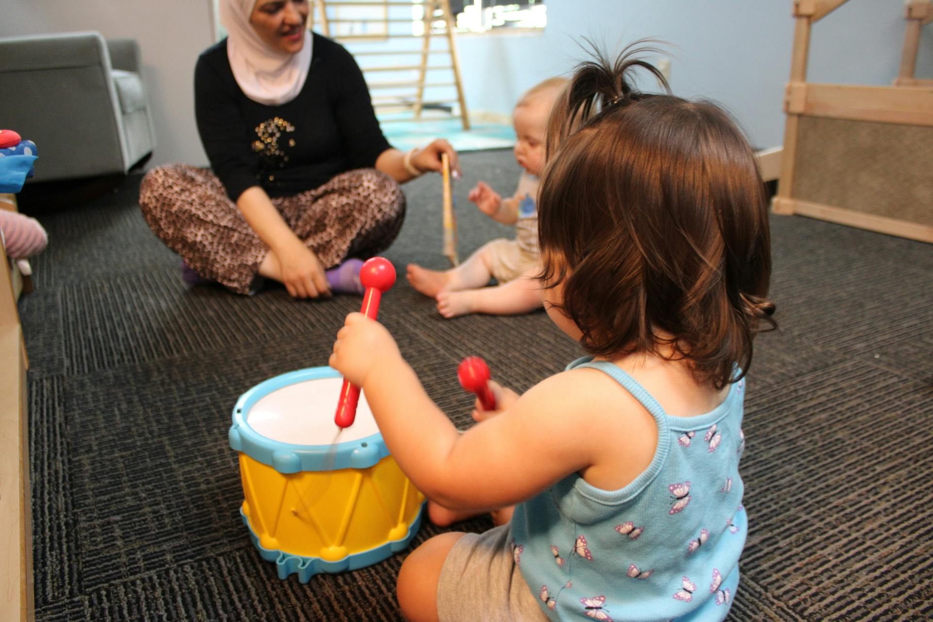 A child hitting a toy drum with mallets while another child and adult watch