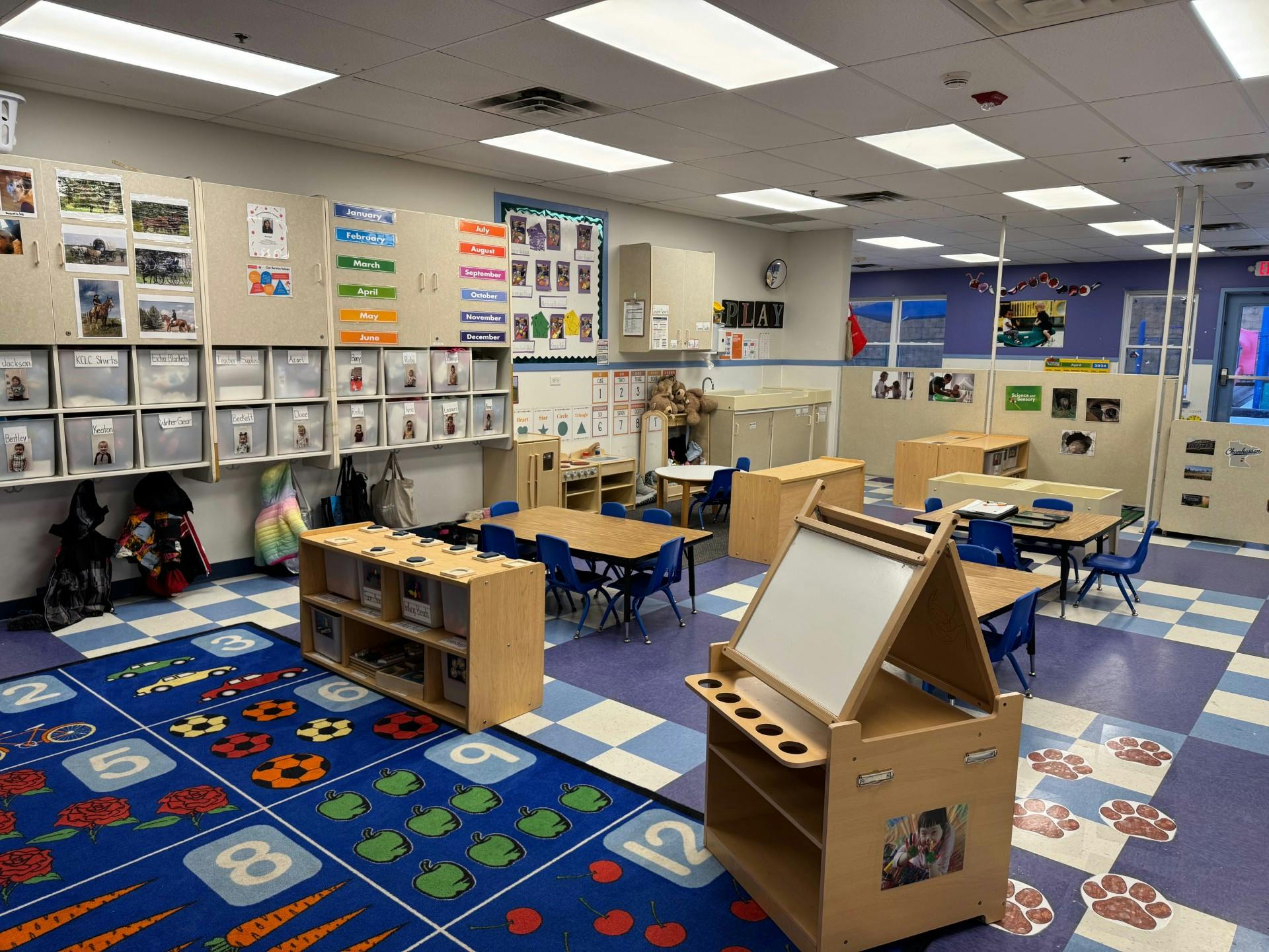 Classroom with small tables, chairs, shelving, and a colorful rug with numbers