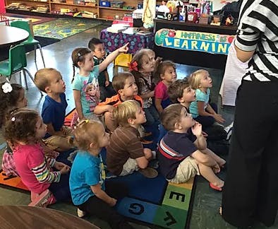 Group of children sitting on a rug looking up at a person