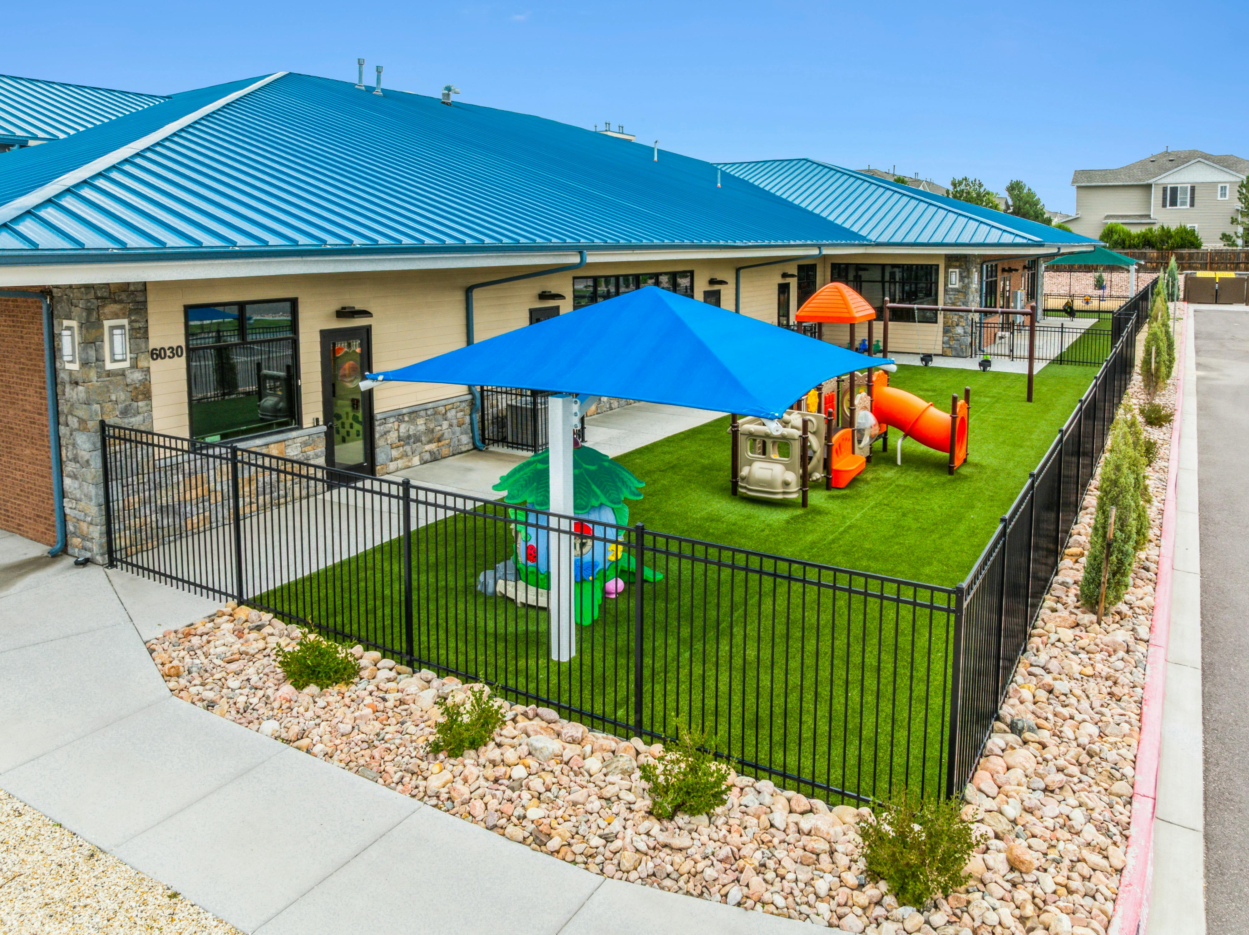 A low-rise building with a blue roof, and an enclosed green turf playground