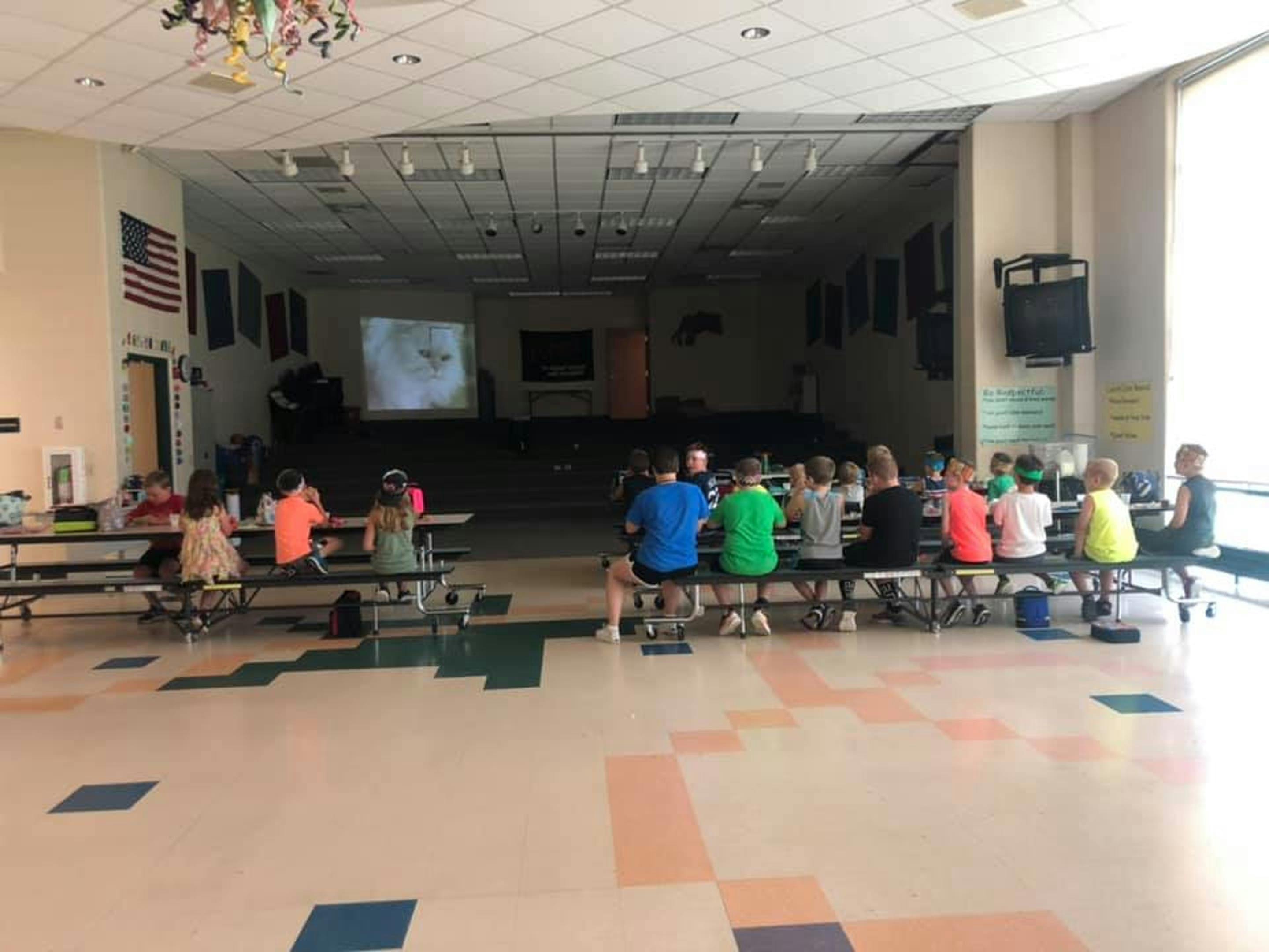 Children sitting at tables in a large room with a projected image