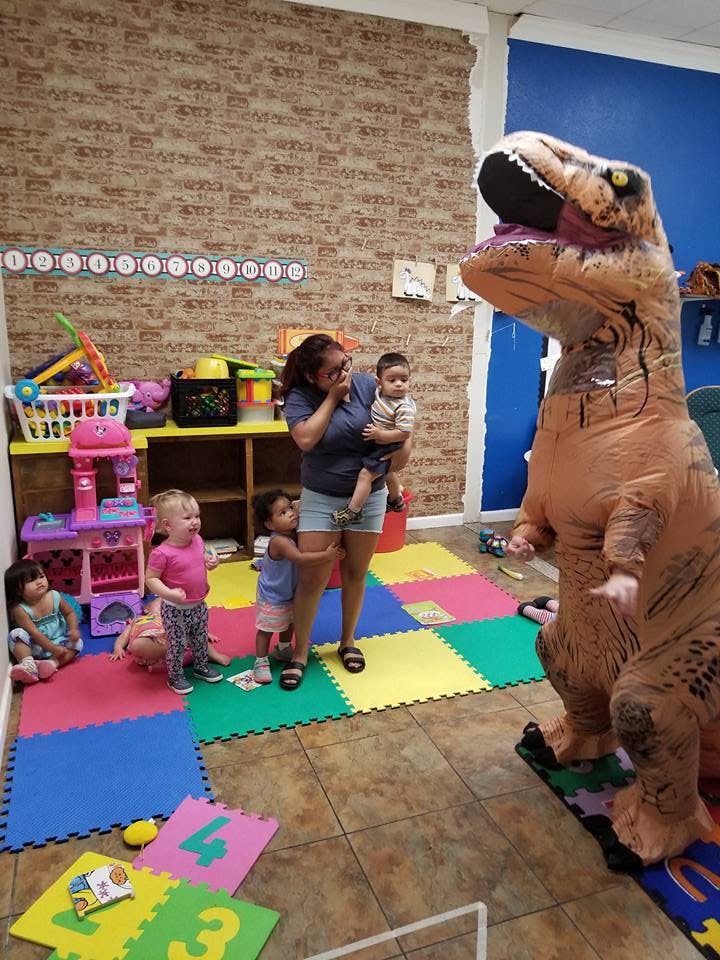 Person holding a child, with other children and a person in a dinosaur costume