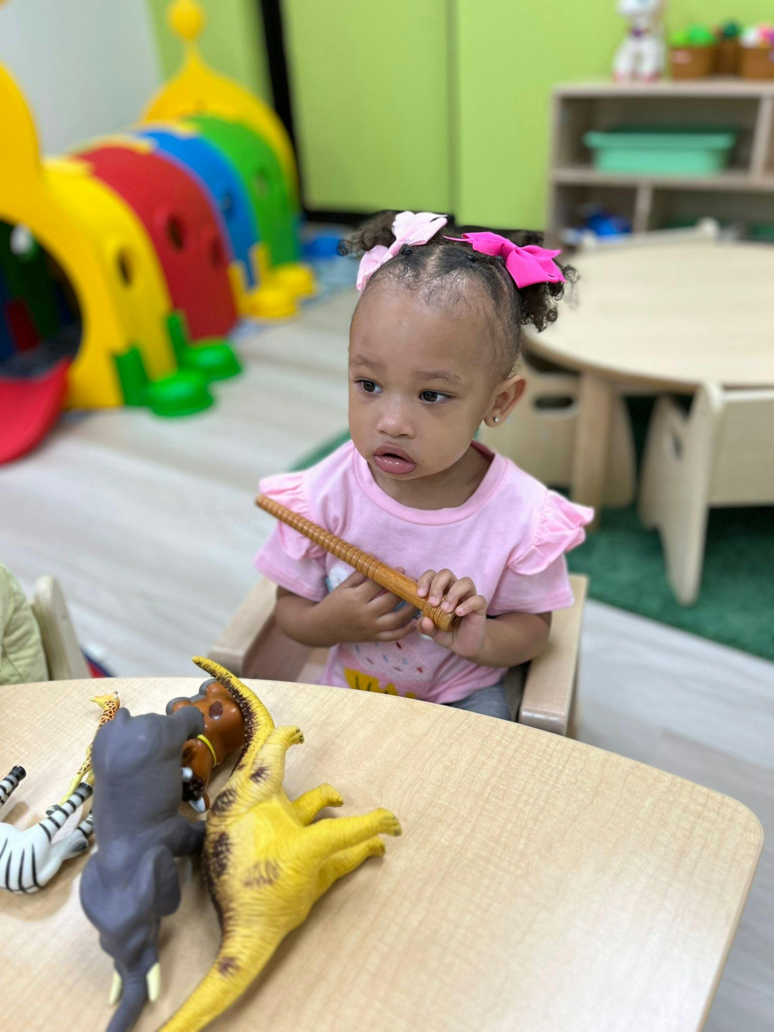 Child sitting at a table with toy animals, holding a brown stick