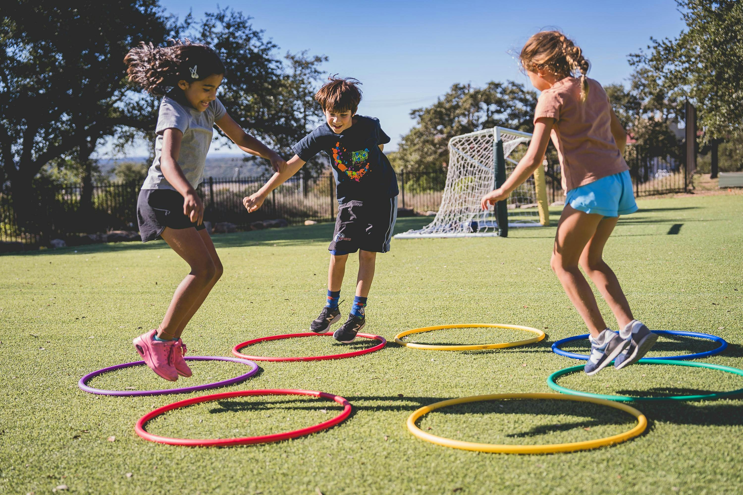 Three children playing on a grass field, jumping over colorful hoops