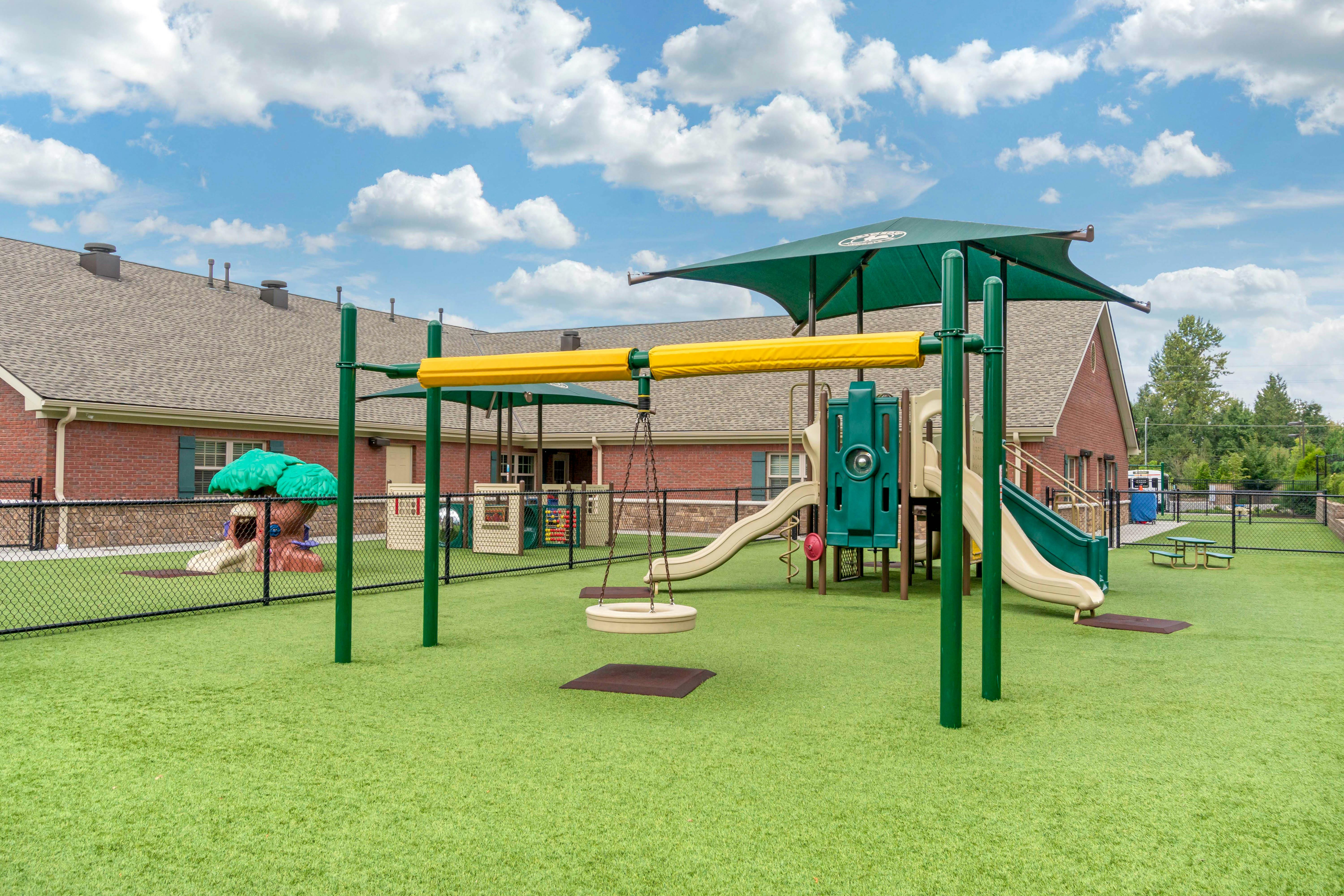 Playground with swings, slides, and green turf in front of a brick building