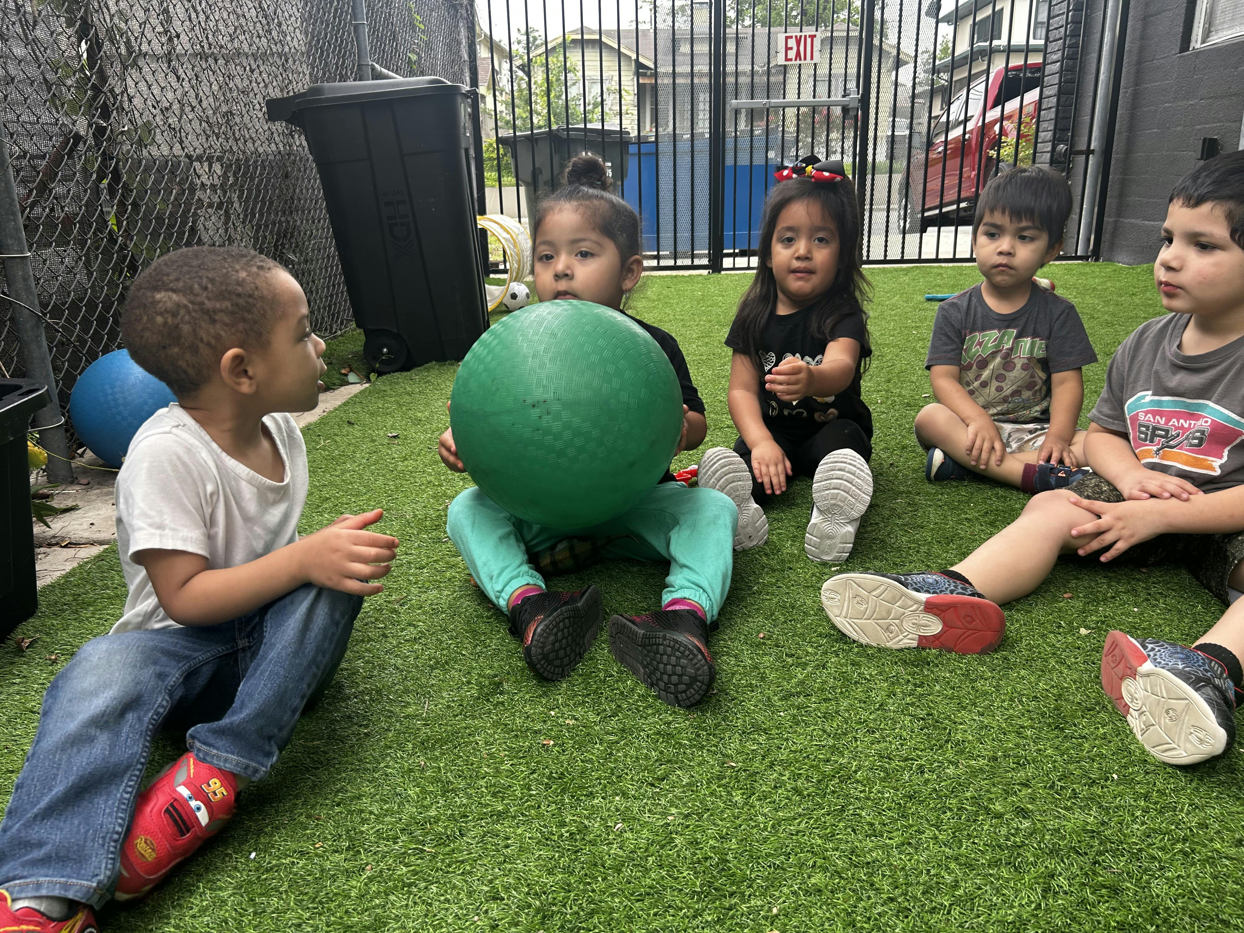 Five children sitting on artificial grass, one child holding a green ball