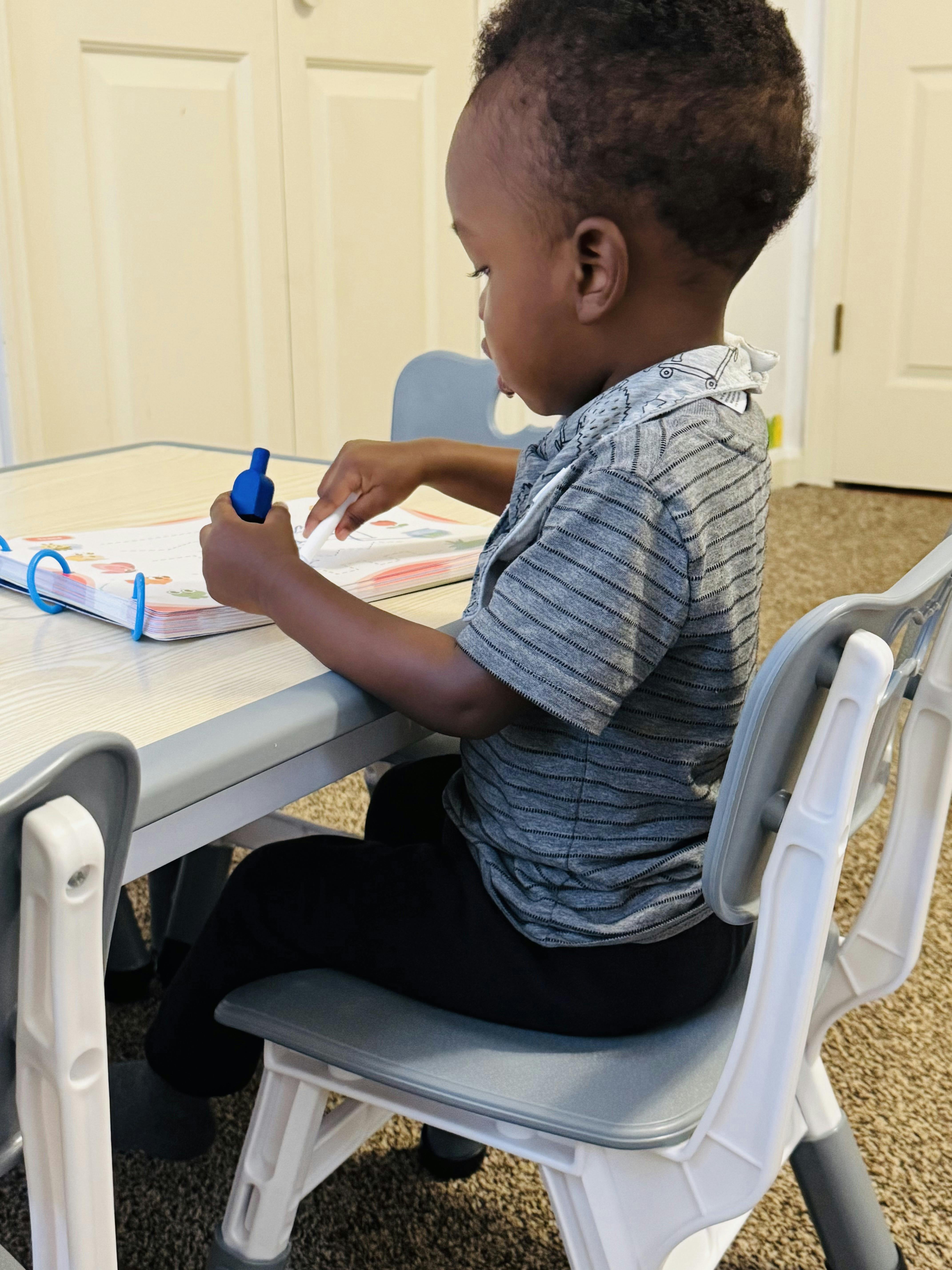 Child sitting at a table, holding a blue marker and looking at a book