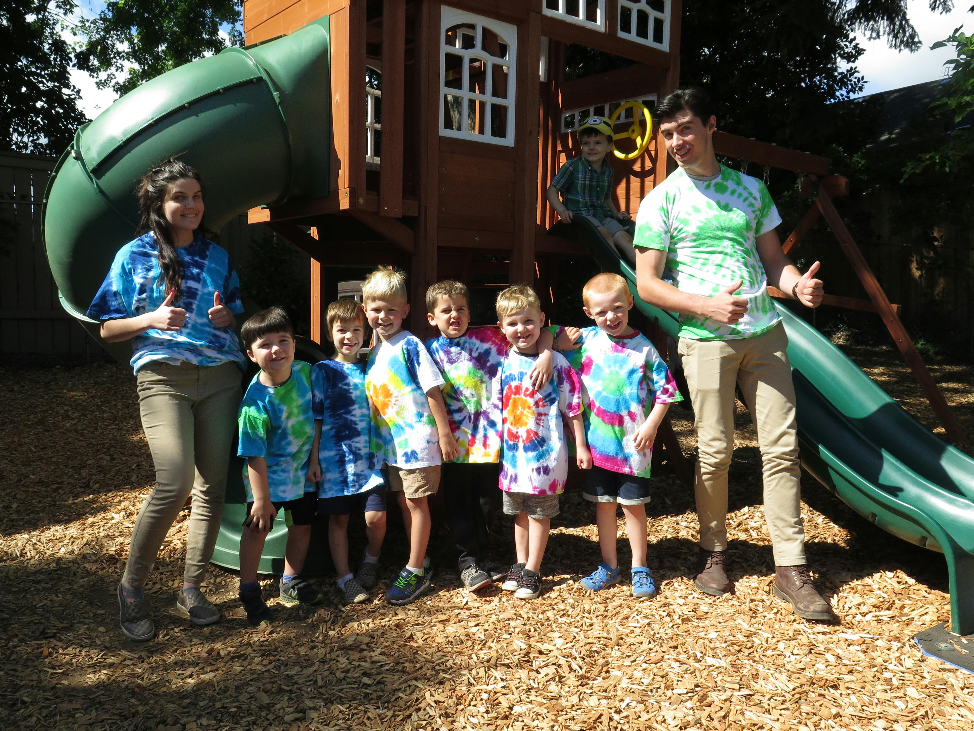 Two people and multiple children in tie-dye shirts standing by a playground slide