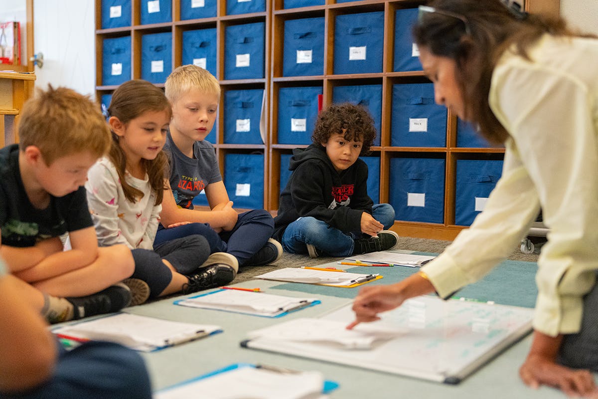 Adult pointing to a whiteboard with children sitting on the floor in a classroom