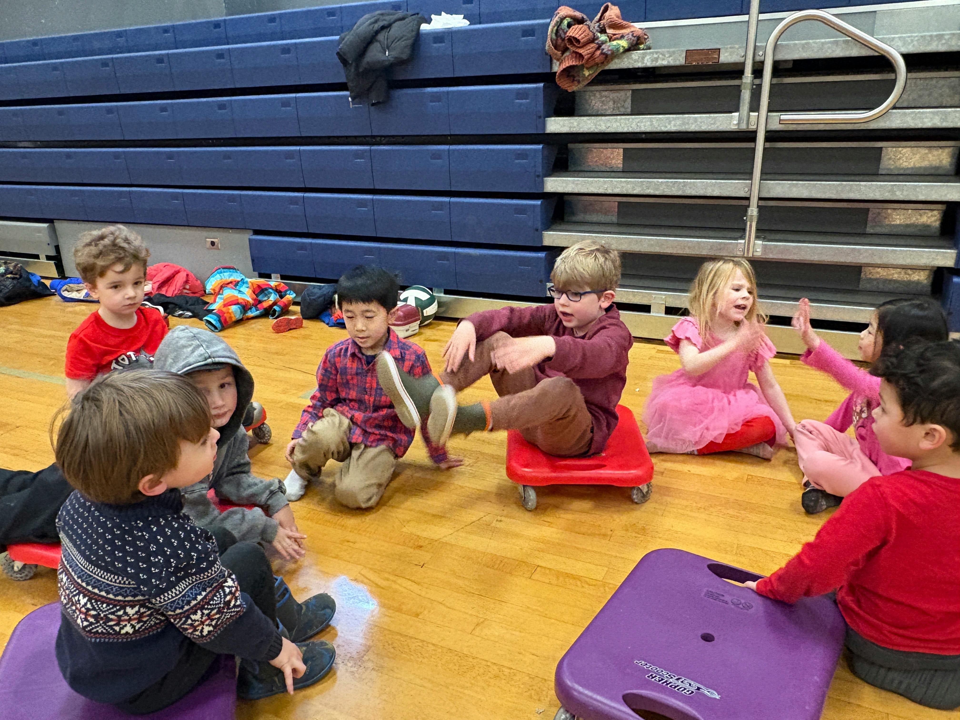 Children sitting on the floor and on scooter boards in a gym
