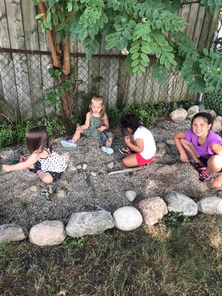 Three children playing in a dirt area with rocks and a tree