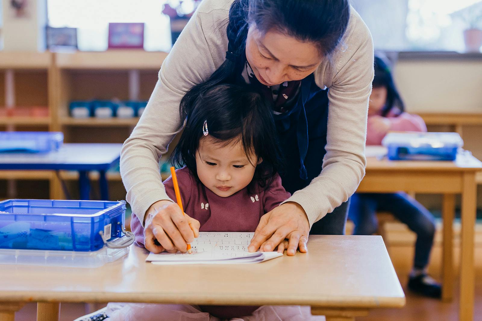 Adult helping a child write at a desk in a classroom