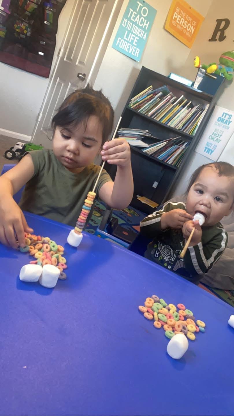 Two children threading cereal onto skewers at a table