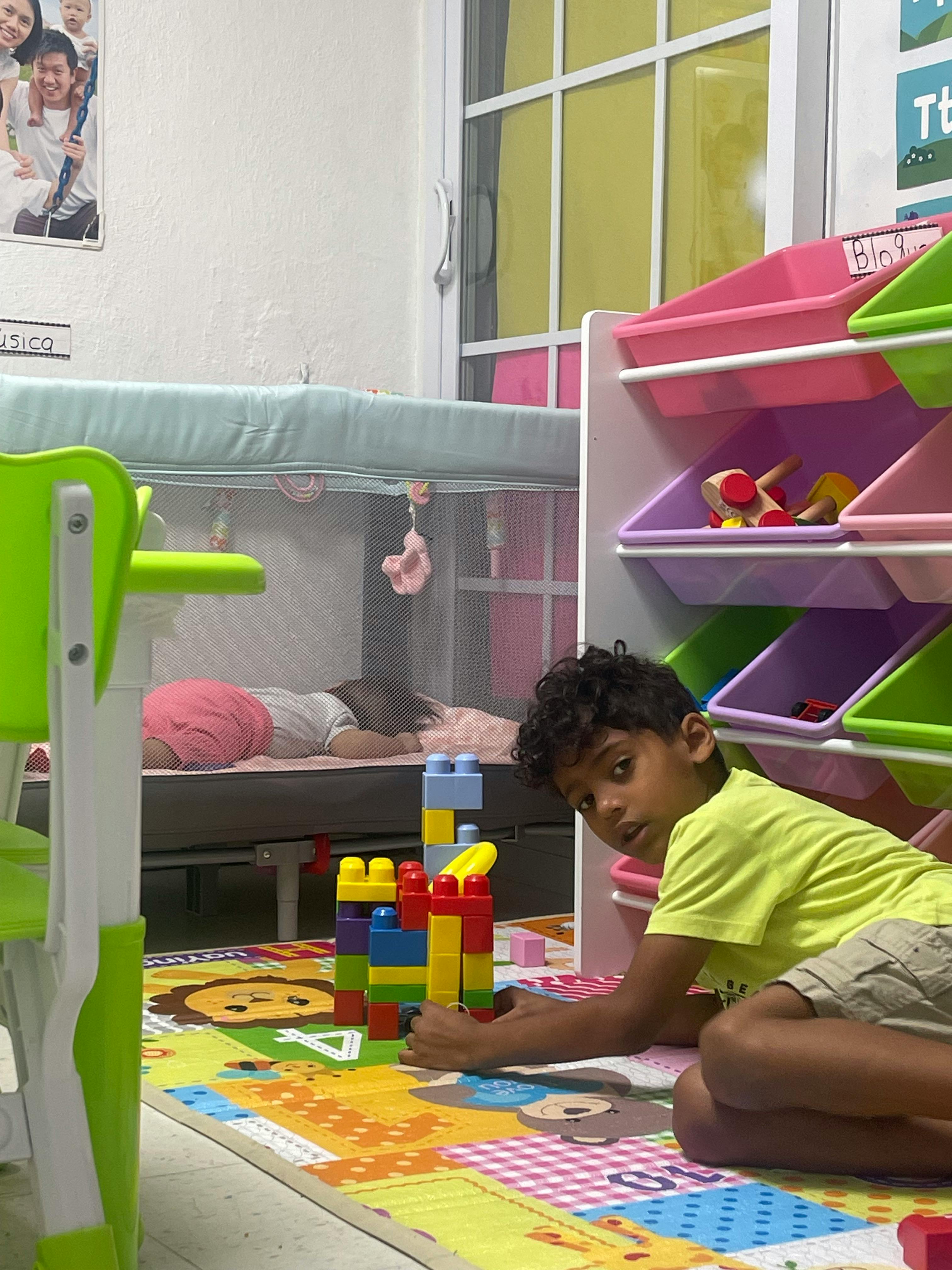 Child playing with building blocks on a mat, with another child sleeping in a crib