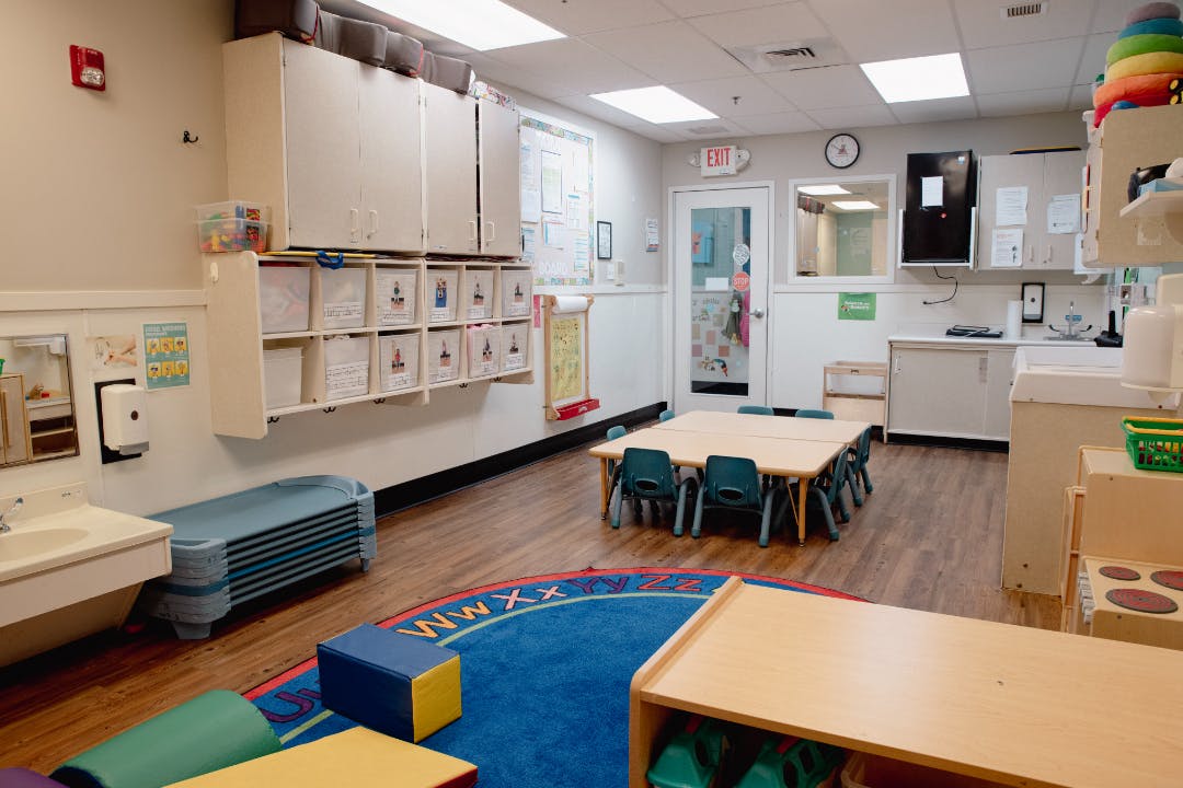An empty children's classroom with small tables and chairs, storage, and a sink area
