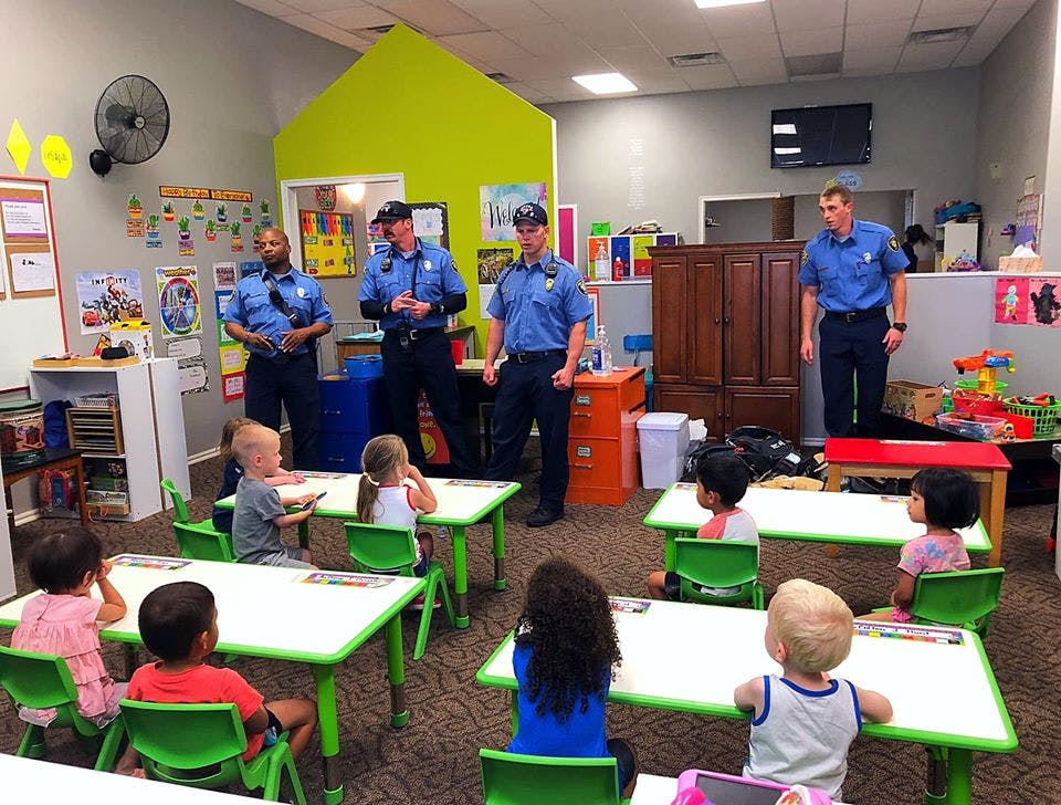 Four people in uniform standing in a classroom with children seated at desks