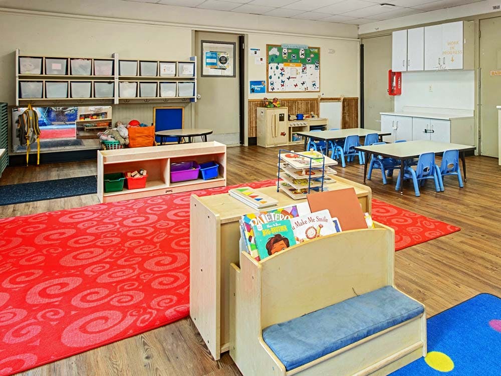 A classroom with tables, chairs, storage cubbies, and a bookshelf with books