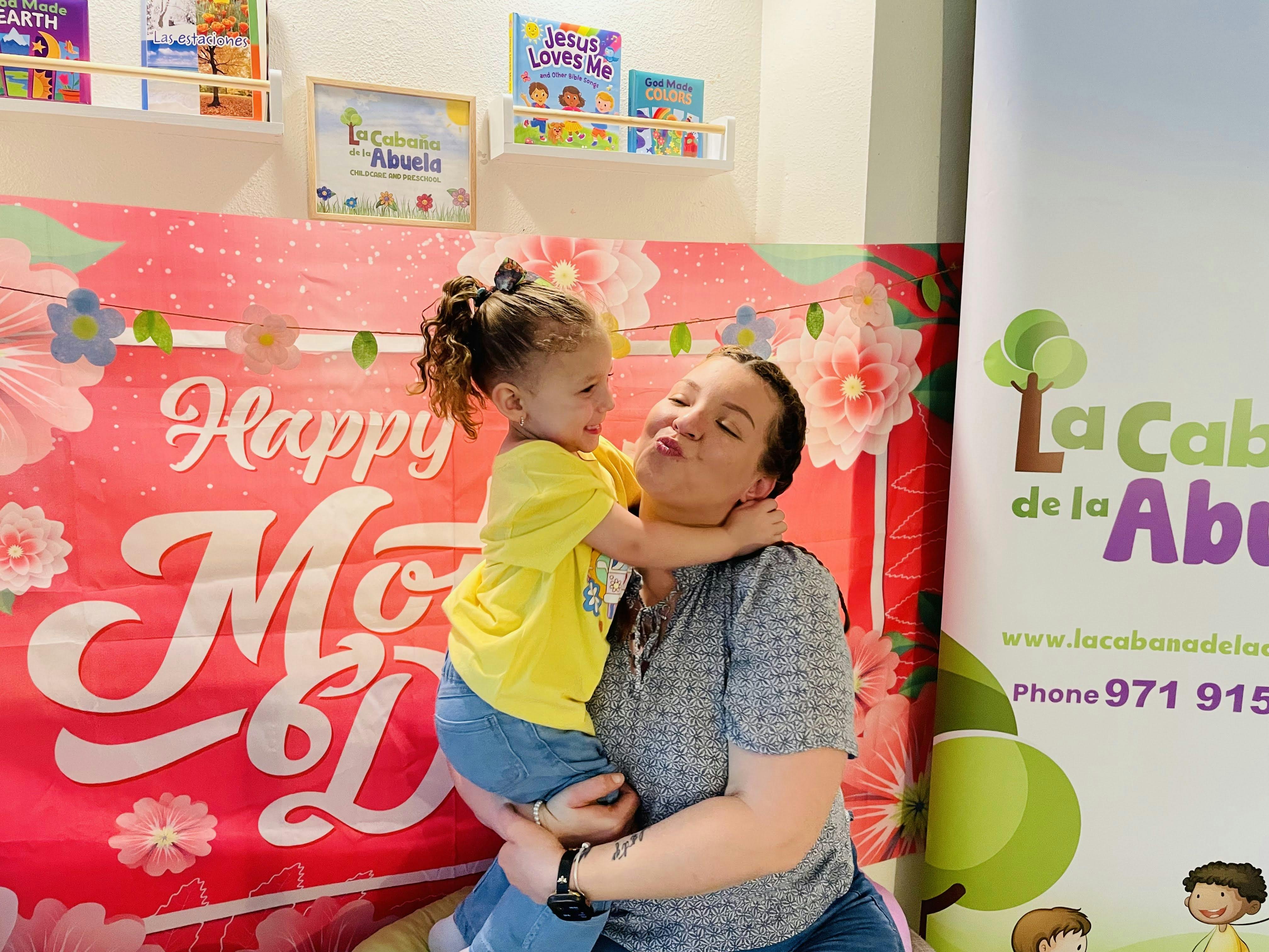 Adult holding a child in front of a Happy Mother Day backdrop
