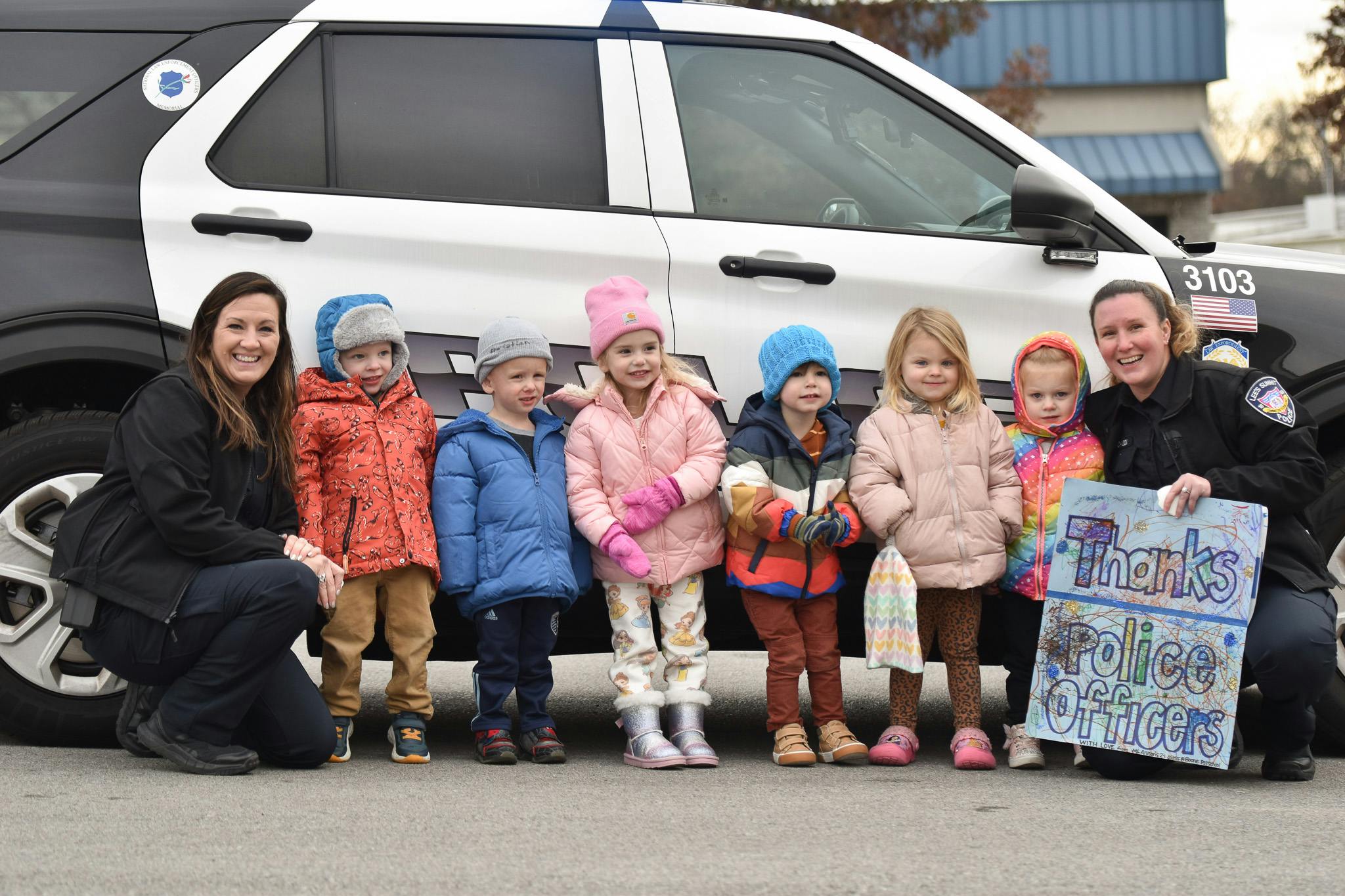 Two people and six children in winter clothing standing in front of a police car, holding a sign that reads "Thanks Police Officers"