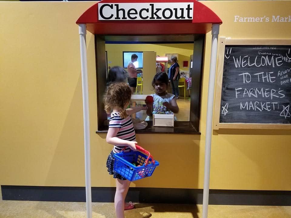 Child holding a blue basket at a checkout counter with another child inside
