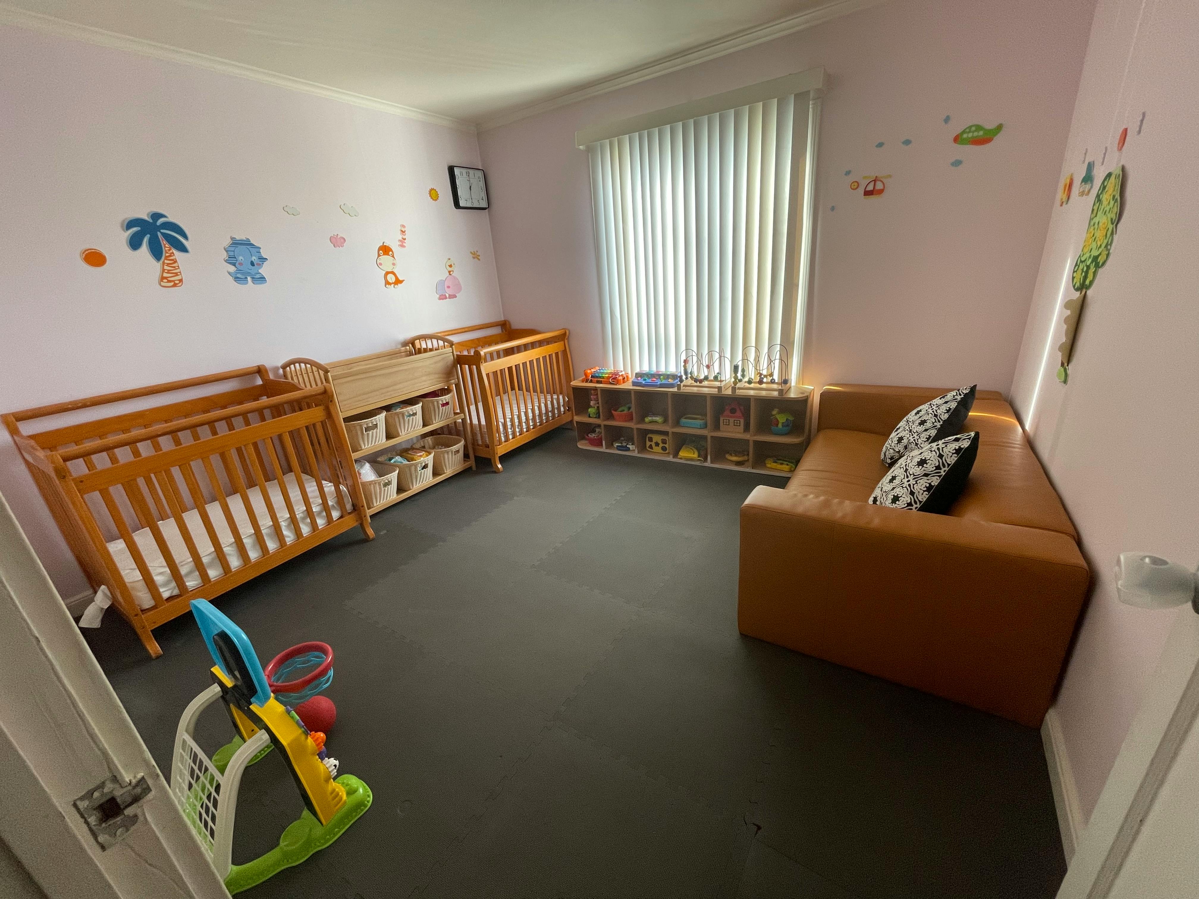 Nursery room with two cribs, a brown sofa, and a window with blinds