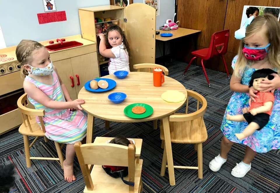 Three children playing in a classroom, one holding a doll