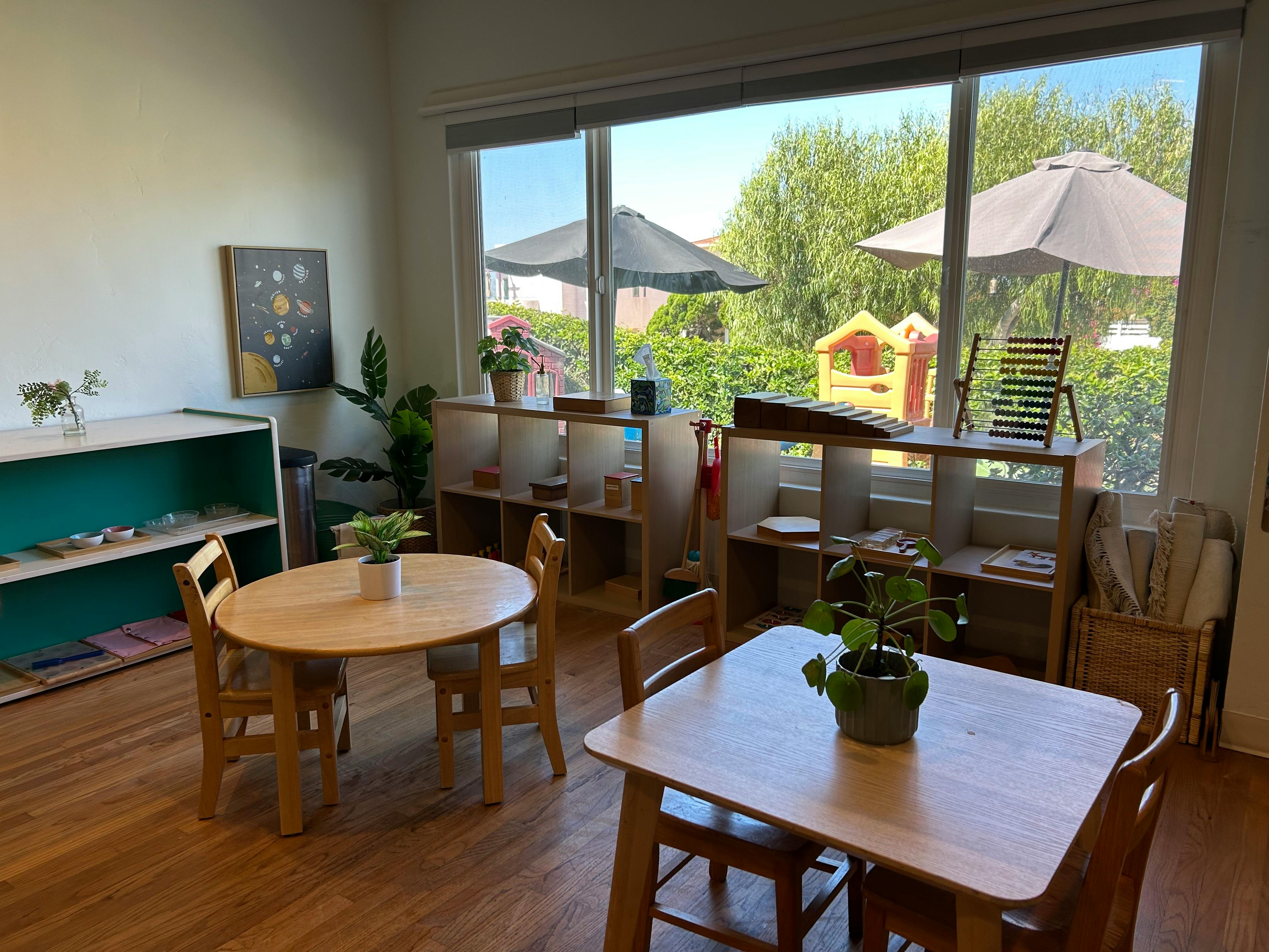 Classroom with tables and chairs, shelves, and large windows overlooking a yard
