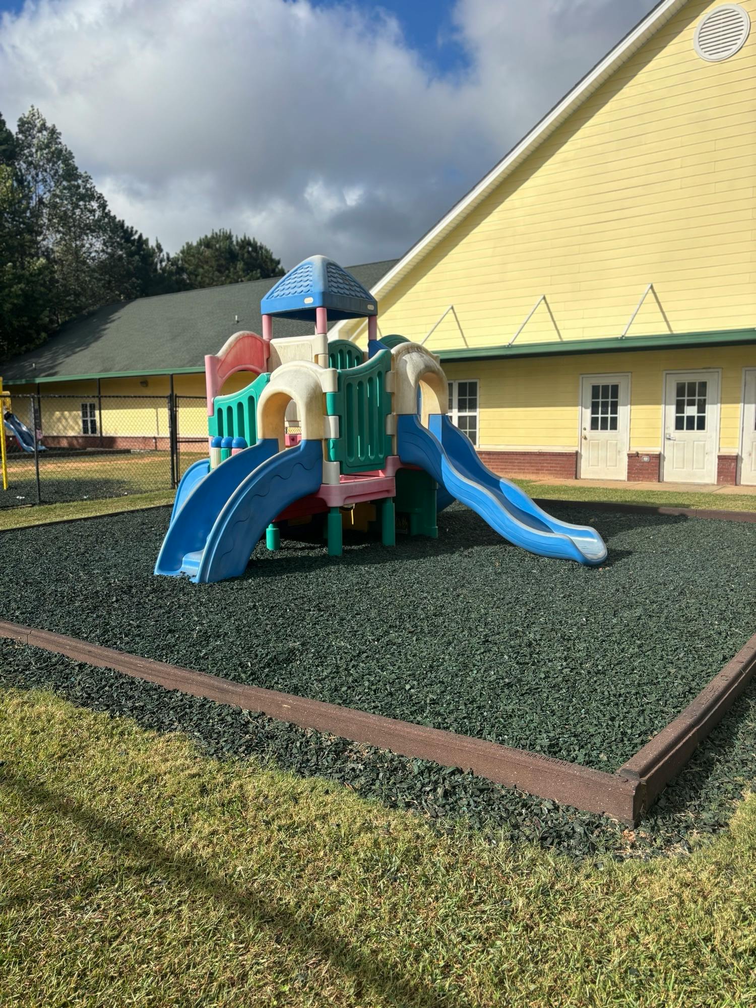 A colorful playground structure with slides on rubber mulch next to a yellow building