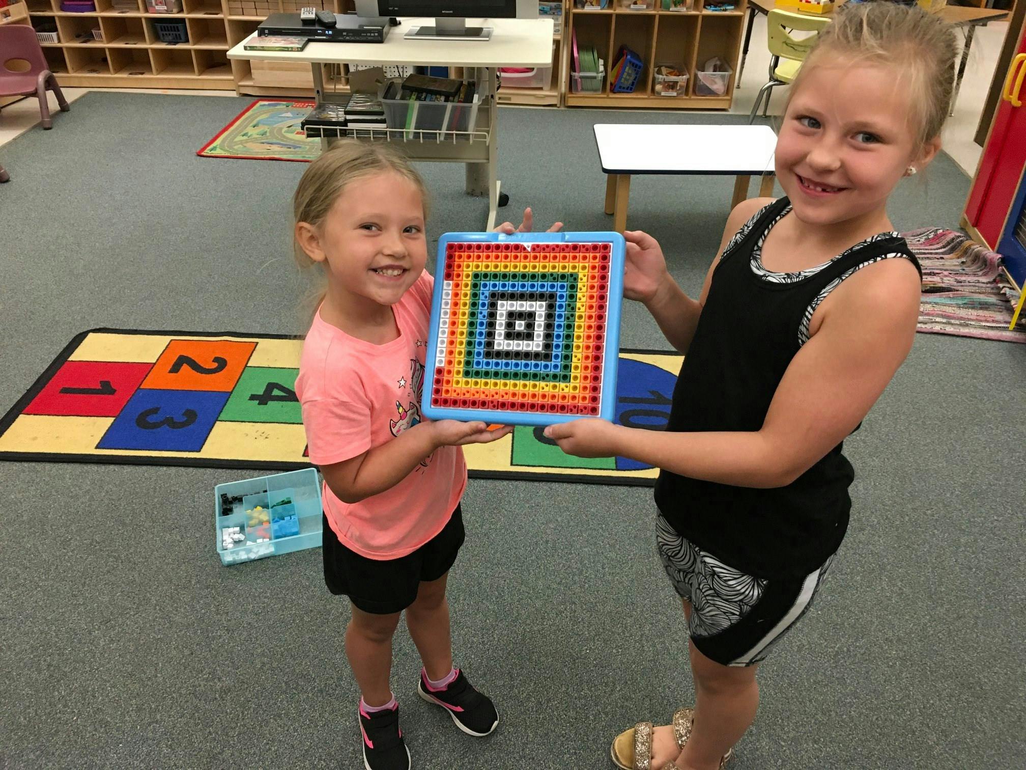 Two children holding up a colorful square pegboard design in a classroom