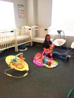 Person sitting on floor with child, baby bouncer and play mat, cribs in background
