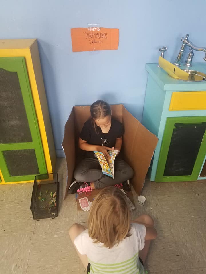 Child sitting in a cardboard box reading a book, another child sitting nearby