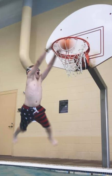 Child jumping to dunk a basketball into a hoop over a swimming pool