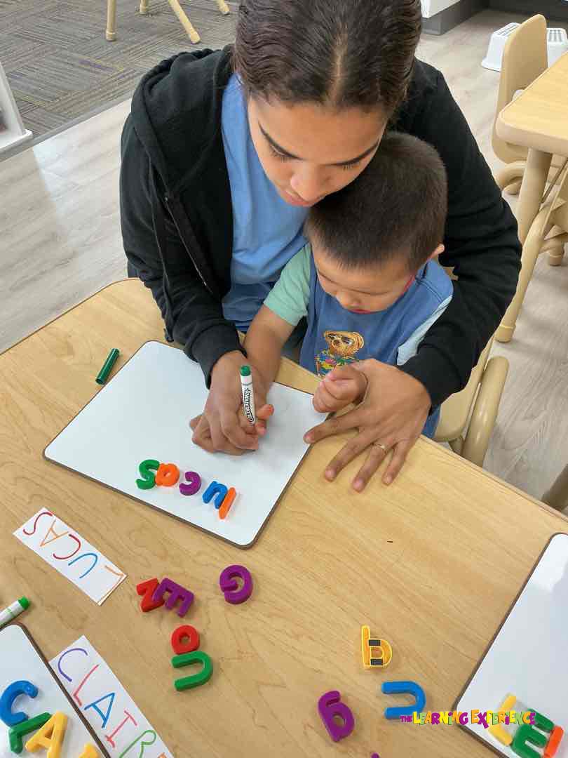 Person helping a child write on a whiteboard with letter magnets