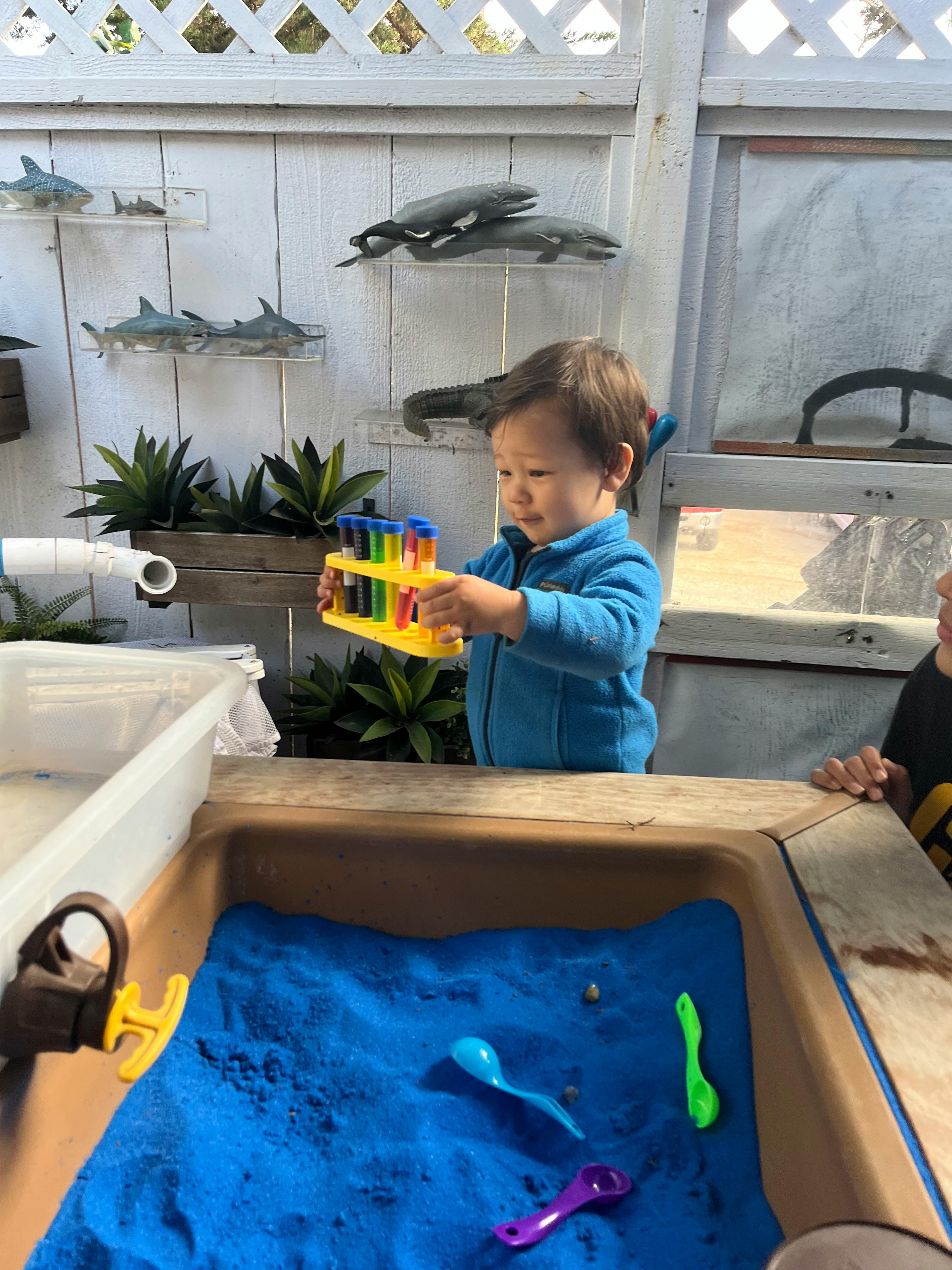 Child holding a toy test tube rack over a sand table